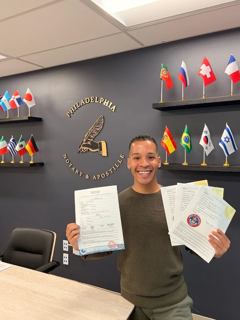 Brian Perez of Philadelphia Notary & Apostille holding apostilled documents in front of the firm's office wall with international flags